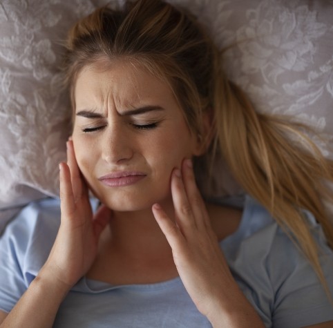 Woman in bed holding her cheeks in pain needing nightguard for bruxism
