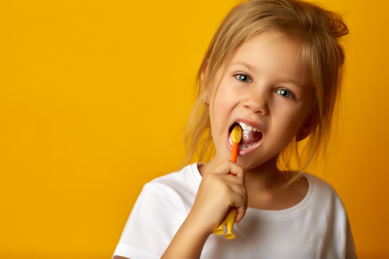 a little girl wearing a white blouse and brushing her teeth with a manual toothbrush